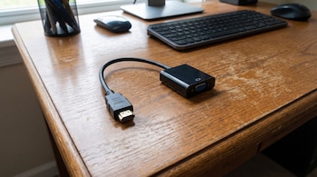 Primer plano de un adaptador de video negro con conectores HDMI y VGA sobre una superficie de escritorio de madera, con un teclado y ratón desenfocados al fondo.