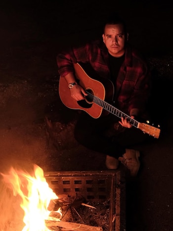 Hombre joven con camisa a cuadros roja y negra toca una guitarra acústica frente a una fogata en la oscuridad, mirando a la cámara