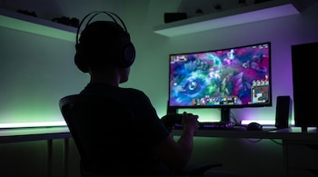 Silueta de una persona con auriculares y gamepad jugando en un monitor curvo, con luces LED verdes y púrpuras en el entorno oscuro de la habitación.