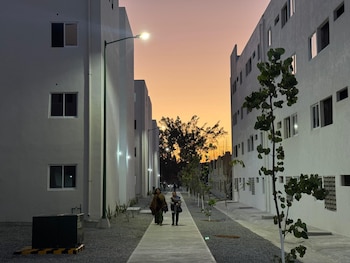 Dos mujeres caminan por un sendero de cemento entre edificios residenciales de varias plantas en Chiapas. Farolas encendidas y un cielo anaranjado al atardecer