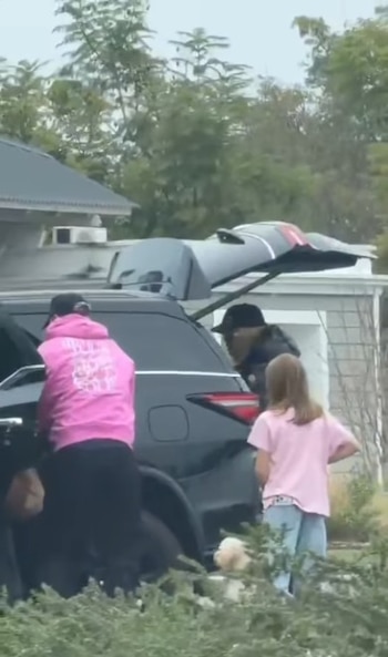 Personas descargando equipaje de una SUV negra. Una mujer en sudadera rosa, un hombre y una niña en camiseta rosa están cerca del vehículo