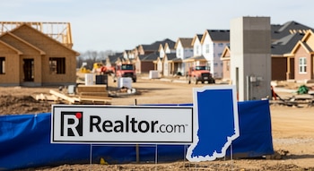 Un cartel de Realtor.com y una silueta azul del estado de Indiana en primer plano, con un gran sitio de construcción de casas en un suburbio al fondo.