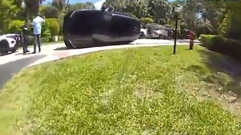 Un vehículo negro volcado de lado sobre un camino pavimentado junto a césped verde. Se ven personas y árboles borrosos en el fondo de la escena