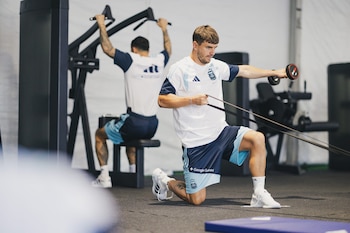 Dos futbolistas con indumentaria oficial azul y blanca entrenan en un gimnasio. Uno arrodillado con una banda de resistencia, el otro sentado en una máquina