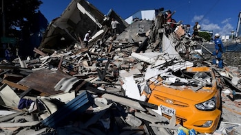 Montones de escombros de un edificio destruido, con personas sobre ellos y un coche naranja aplastado debajo, bajo un cielo azul en Pereira, Colombia