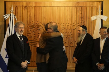 Cinco hombres en un salón con una pared de madera. Dos hombres se abrazan. Se observa una bandera de Israel y dos decoraciones blancas en forma de lazo
