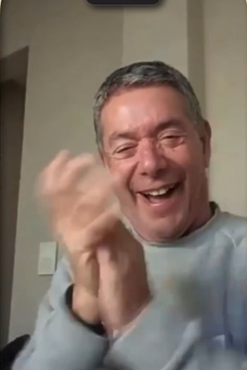 Un hombre de mediana edad, con cabello gris, ríe con la boca abierta mientras agita sus manos en primer plano, viste una sudadera gris