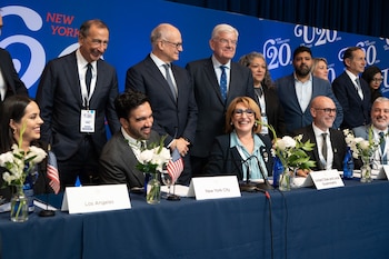 Personas en traje sentadas y de pie en una mesa de conferencia con flores, banderas de Estados Unidos y letreros de ciudades ante un fondo azul