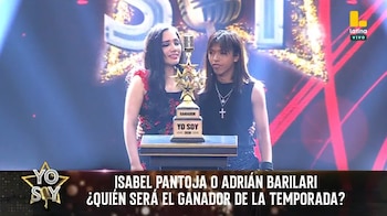 Dos personas de pie junto a un trofeo dorado con la inscripción Yo Soy sobre el escenario iluminado de un estudio de televisión