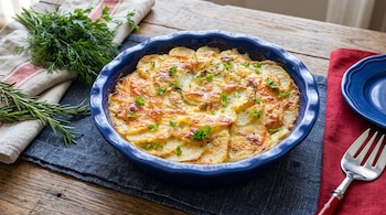 Un molde azul con pastel de patata y queso gratinado, junto a hierbas frescas, un tenedor y un plato pequeño sobre una mesa de madera.