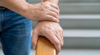 Primer plano de dos manos de adulto con vello corporal, una con anillo de oro, sobre un pasamanos de madera; visibles pantalones de mezclilla y escaleras difusas.