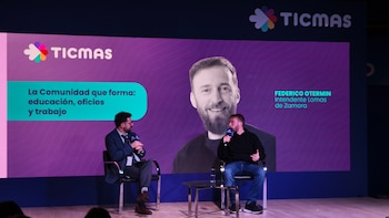 Dos hombres sentados en sillas en un escenario frente a una gran pantalla con el logo de TICMAS y una imagen de José Joaquín Brunner; uno sostiene un micrófono