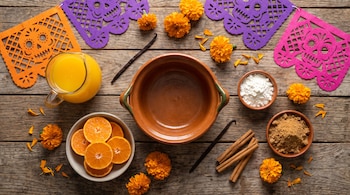 Vista cenital de mandarinas, jugo, fécula de maíz, azúcar, canela, una olla de barro, flores de cempasúchil y papel picado sobre madera.
