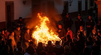 Multitud de personas alrededor de una gran hoguera en una calle. Tres agentes de policía uniformados observan detrás de la gente. Edificios de noche al fondo.