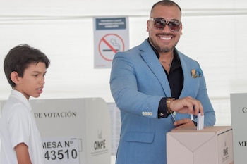 Hombre sonriente con traje azul y gafas de sol deposita un voto en una urna electoral. Un niño lo mira a su izquierda. Al fondo, una cabina de votación y un cartel de no fumar