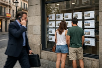 Una pareja de espaldas mira un escaparate de inmobiliaria con anuncios de propiedades; un hombre con traje y maletín pasa hablando por teléfono.