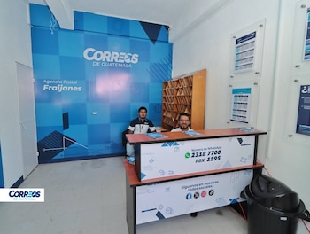 Dos hombres detrás de un mostrador en una agencia postal. Fondo con banner azul de "Correos de Guatemala" y "Agencia Postal Fraijanes", casilleros y avisos
