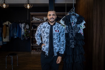 Un hombre sonriente en una tienda de ropa. Viste chaqueta estampada y pantalón oscuro. A su alrededor hay estantes con ropa, un perchero y lámparas