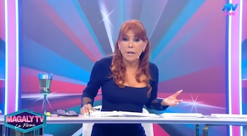 Magaly Medina sentada en un escritorio en un estudio de televisión con fondo azul y rosa, usando un vestido azul oscuro, hablando y gesticulando. Hay documentos, lentes y una copa sobre la mesa