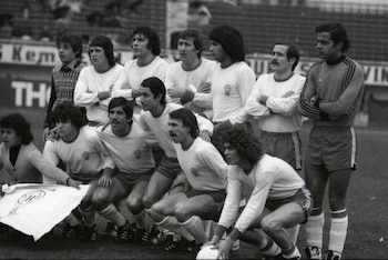 Doce personas, la mayoría con uniformes de fútbol blancos con escudo, posan en un campo de césped. Un arquero usa camiseta a rayas. Un balón en el suelo