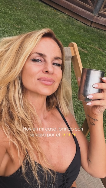 Una mujer rubia sonríe mientras sostiene un vaso metálico en una silla de jardín, con césped al fondo y texto en pantalla