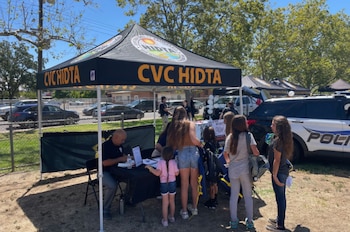 Una carpa negra con la inscripción CVC HIDTA cobija una mesa de atención a varias personas, junto a una patrulla policial al aire libre