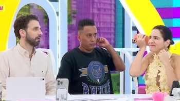 Tres personas en un estudio de televisión. Christian Cueva, en el centro, toca su oreja. A la derecha, una mujer sonríe y sostiene un teléfono. A la izquierda, un hombre observa sorprendido