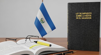Libro negro con 'LEY DE IMPUESTO SOBRE LA RENTA DE EL SALVADOR' en oro, libro abierto con resaltador, gafas, pluma y una bandera salvadoreña en una mesa de madera.