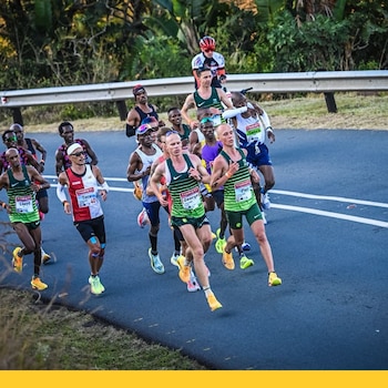 Grupo de corredores varones en una carretera. Visten camisetas deportivas y dorsales. Se observa un quitamiedos, vegetación y un ciclista al fondo