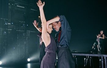 Dos bailarines en un escenario oscuro. La mujer de negro tiene los brazos levantados. El hombre de camisa oscura y tirantes rojos la abraza por la cabeza. Focos en el fondo