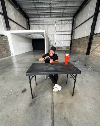 Nicolás Magaldi sonriente, con gorra y camiseta negra, sentado en una mesa oscura preparando mate en un gran galpón con paredes de ladrillo y chapa metálica