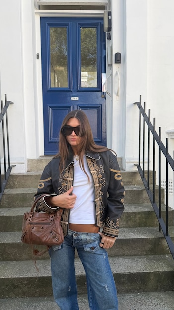 Lola Latorre de pie en escalones de piedra frente a puerta doble azul. Viste chaqueta negra bordada en oro, camiseta blanca, jeans y gafas de sol