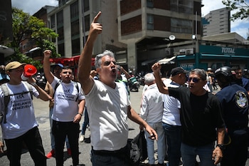 Un hombre grita y señala con un dedo, otro grita con un puño en alto. Un manifestante usa un megáfono y viste una camiseta del Movimiento Adelante Venezuela. Hay un oficial de policía