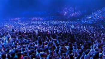 Vista aérea de una enorme multitud de personas en un concierto, iluminadas por luces azules intensas. Se ven cabezas y brazos levantados