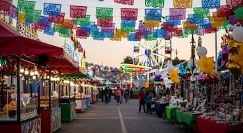 Corredor de feria mexicana con banderines de papel picado, puestos de comida, puestos de artesanías, globos y juegos mecánicos iluminados al fondo.