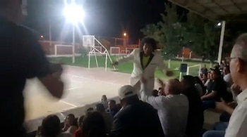 Un hombre con traje blanco y pelo rizado en el centro de un escenario al aire libre, bajo intensas luces nocturnas, rodeado por una multitud difusa.