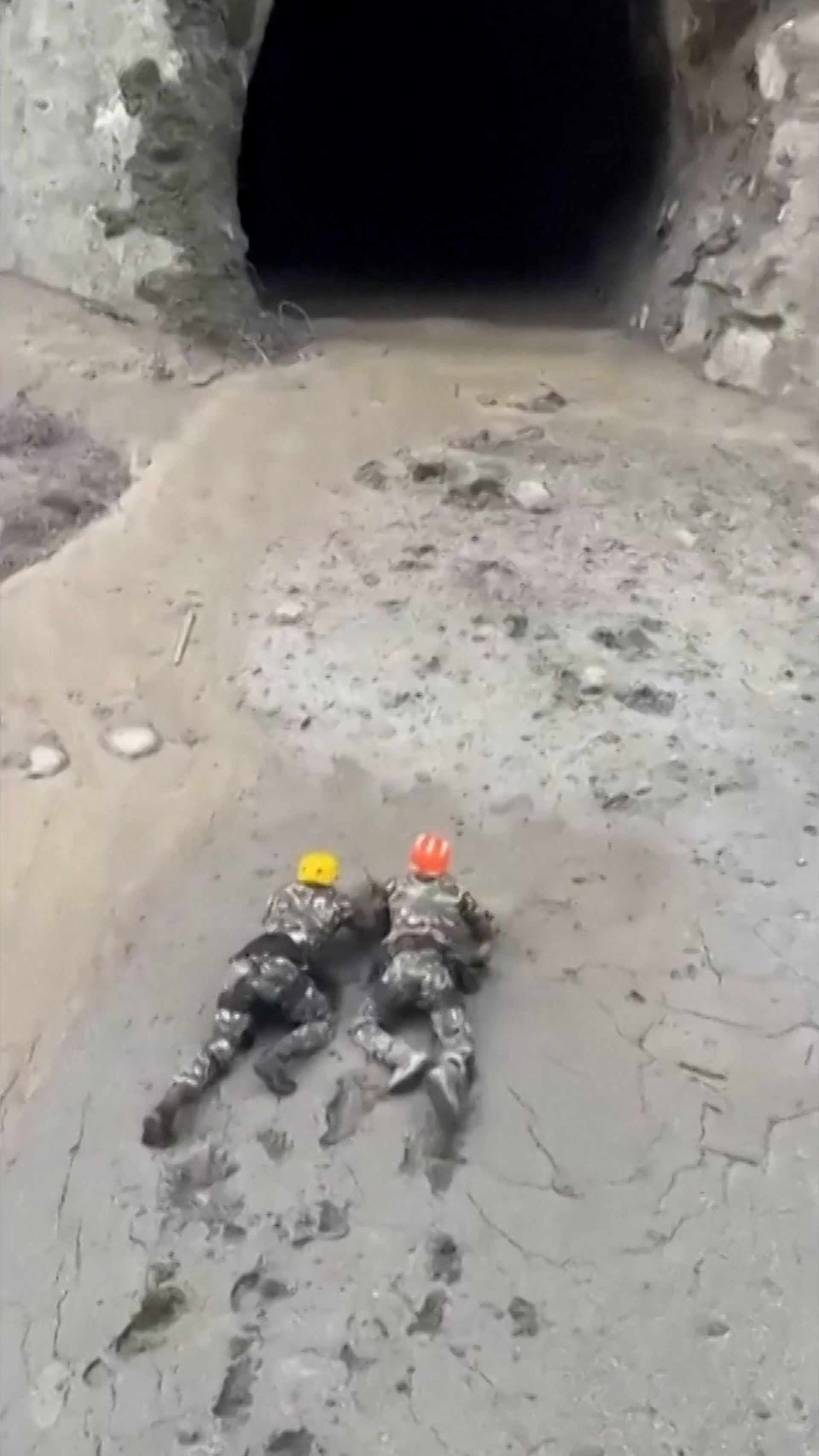 Dos rescatistas con cascos de seguridad se arrastran por el lodo hacia una entrada oscura durante las labores de salvamento en Nepal (Captura de video)