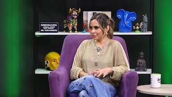 Una mujer con blusa dorada y pantalón de mezclilla sentada en un sillón morado delante de un estante con figuras decorativas