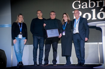 Cinco personas posan en un escenario con fondo oscuro y un texto blanco en la pantalla. El hombre del centro sostiene un premio o placa conmemorativa