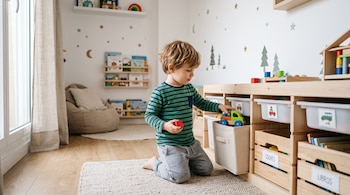 Un niño arrodillado guarda juguetes de madera en estantes etiquetados dentro de una habitación iluminada.