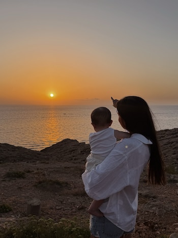 Mujer de espaldas con un bebé en brazos señalando el sol poniente sobre el mar. El horizonte muestra un atardecer naranja
