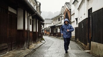 Un hombre con chaqueta azul con capucha camina por una calle empedrada mojada entre casas tradicionales con fachadas de madera y yeso blanco.