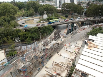Obra en construcción con andamios, trabajadores, un puente vehicular, tráfico, una rotonda con una estatua, árboles y edificios urbanos