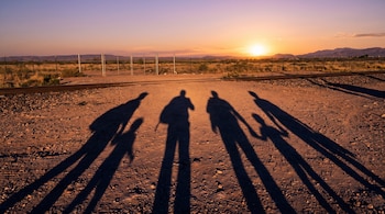 Seis sombras humanas largas sobre un camino de tierra, dos de ellas pequeñas. Al fondo, vías de tren, una valla, vegetación seca, montañas y un atardecer.