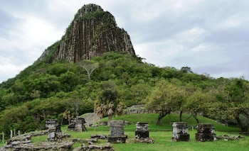Vista de una montaña rocosa con vegetación, ruinas de piedra de un sitio arqueológico y cielo nublado