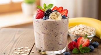 Un vaso de batido de avena con frutos rojos (fresas, frambuesas, moras, arándanos) y rodajas de plátano, adornado con menta. Al fondo, avena y un plátano.