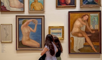 Dos mujeres vistas de espaldas observan cuadros al óleo con desnudos y paisajes colgados en la pared de un museo