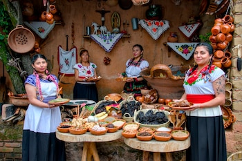 Cuatro mujeres vestidas con blusas bordadas posan junto a mesas de madera con recipientes de barro llenos de especias, chiles secos y vajilla
