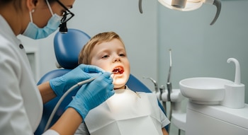Niño pequeño sentado en silla dental azul con babero blanco. Dentista con mascarilla, gafas y guantes azules examina su boca con herramienta iluminada.