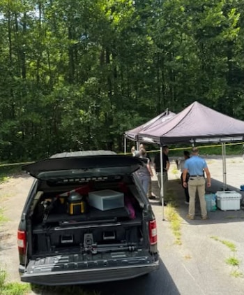 Camioneta pickup negra con la compuerta abierta, dos carpas oscuras, personas, cinta policial amarilla y árboles en un área exterior